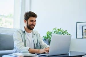 Homem branco sentado no sofá de sua casa mexendo em seu notebook