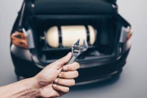Mão segurando chave inglesa em frente a um carro com gnv
